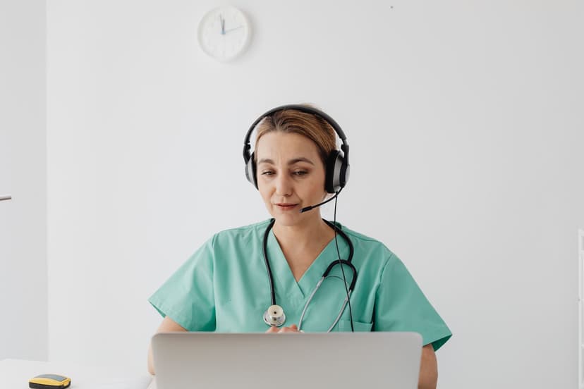 A nurse in green scrubs wearing a headset talks with a patient while working at a laptop.