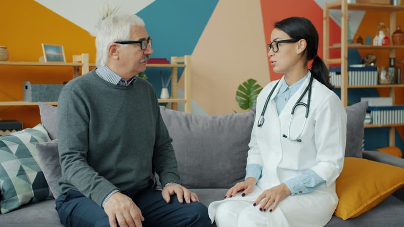 A clinician in a white coat sits on a sofa talking with an older man at home.